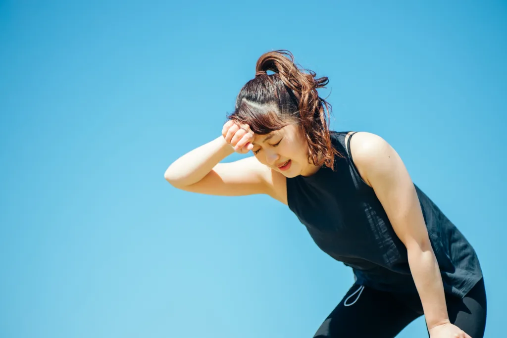 Woman in a black athletic top wipes sweat from her forehead outdoors under a clear blue sky.