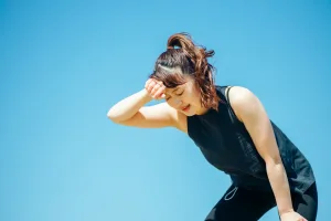 Woman in a black athletic top wipes sweat from her forehead outdoors under a clear blue sky.